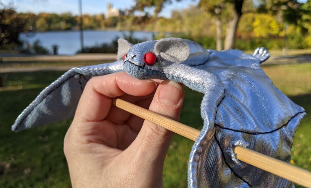a silver bat plush with bright red glass bead eyes and gray ears, a blueish gray cotton underside with a hair stick going thru the wings. this is meant to be worn through a hair bun or ponytail.
