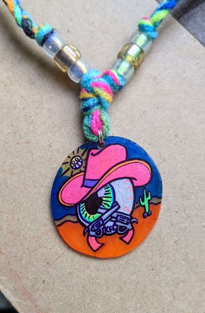 a decent sized dink charm of a green eyeball looking to the side in a noble manner, pink cowboy hat tilted tastefully down while it holds 2 crossed revolvers with pink handles. the background is an orange desert with a blue sky, a cactus, and a very enthusiastic sun saying hiiiiiii

it's hard to tell from this but the background is translucent like stained glass while the foreground and the sun i used gel pens for so they're opaque and slightly textured

the cord is macrame'd yarn with some beads