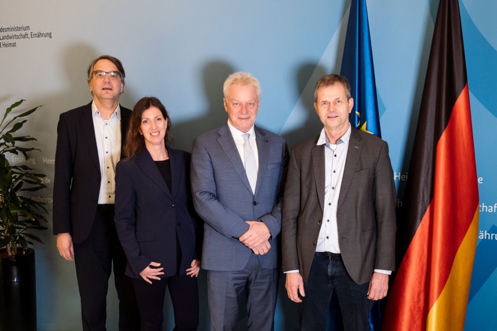 Bundesminister Rainer steht mit drei Personen vor blauem Hintergrund, links eine Pflanze, rechts eine deutsche und EU-Flagge.