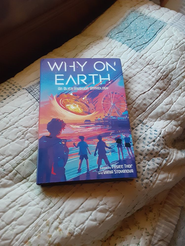 Copy of Why On Earth:An Alien Invasion Anthology on a patchwork quilt. The cover shows a group of people watching a saucer shaped ship crash into the ocean. A pier with a ferris wheel is visible in the foreground and the entire image is rendered in shades of purple, blue, pink, orange, red, and yellow.