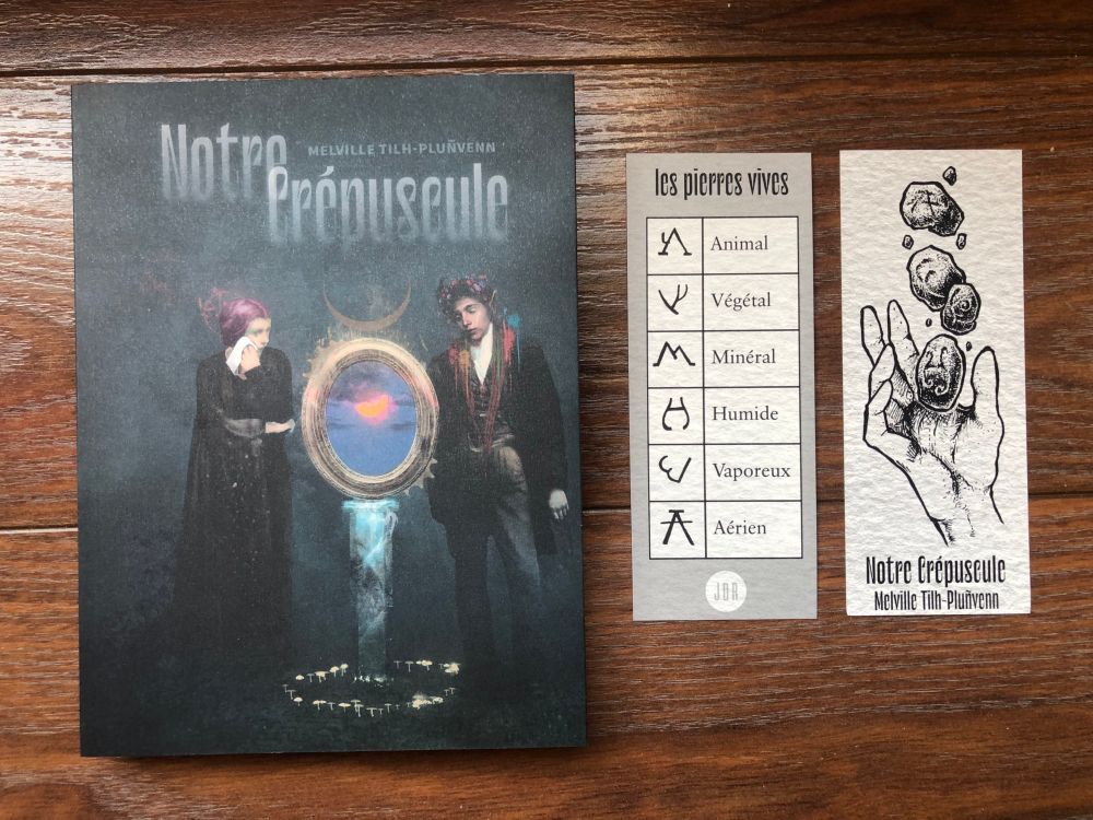 Couverture : deux personnages en deuil devant une stèle où le soleil se couche sous le titre Notre Crépuscule qui s'efface.
Un marque-page : recto la liste des symboles des pierres vives et les éléments auxquels ils renvoient (Animal, Végétal, Minéral, Humide, Vaporeux, Aérien) et au verso une main qui lance des pierres vives.