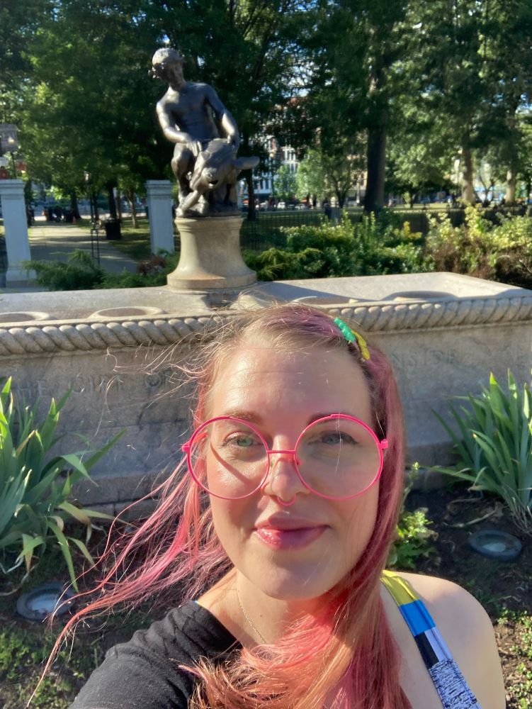 Star St.Germain, a woman with pink hair wearing round pink glasses, standing in front of the Burnside Fountain- a fountain with a bronze statue of a boy riding a turtle.
