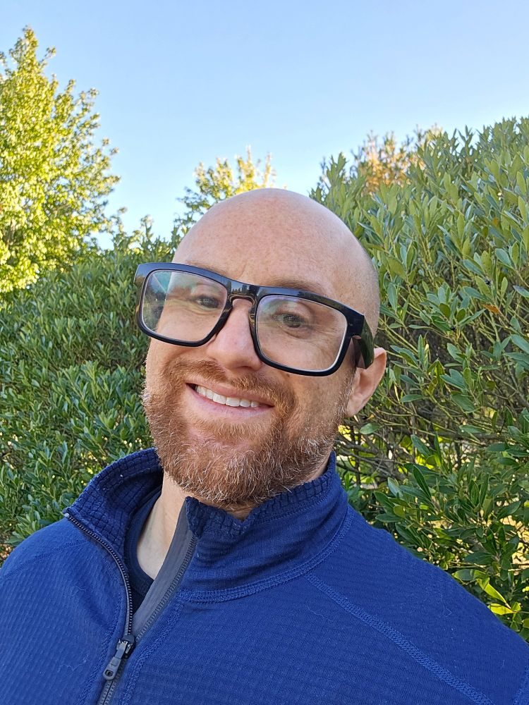 A selfie of me in front of a hedge on a sunny, clear day. I'm a middle-aged, bald, white man with a red beard flecked with white. I'm wearing glasses with thick black rims and a zipped up blue sweater over a black shirt.