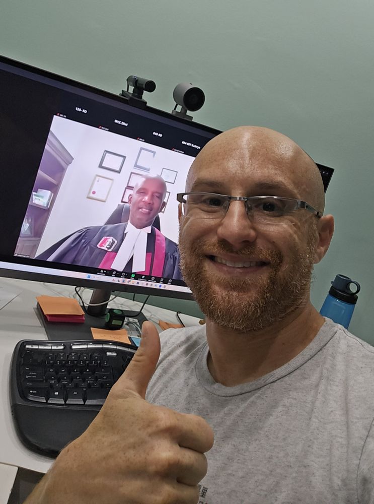 A selfie of me in front of a wide computer monitor with a zoom window open with a robed Canadian judge on it. I'm a bald, middle aged, white man with a red beard flecked with white, giving a thumbs up. I'm wearing glasses with a metal top rim and a grey t-shirt. The judge is a bald black man.