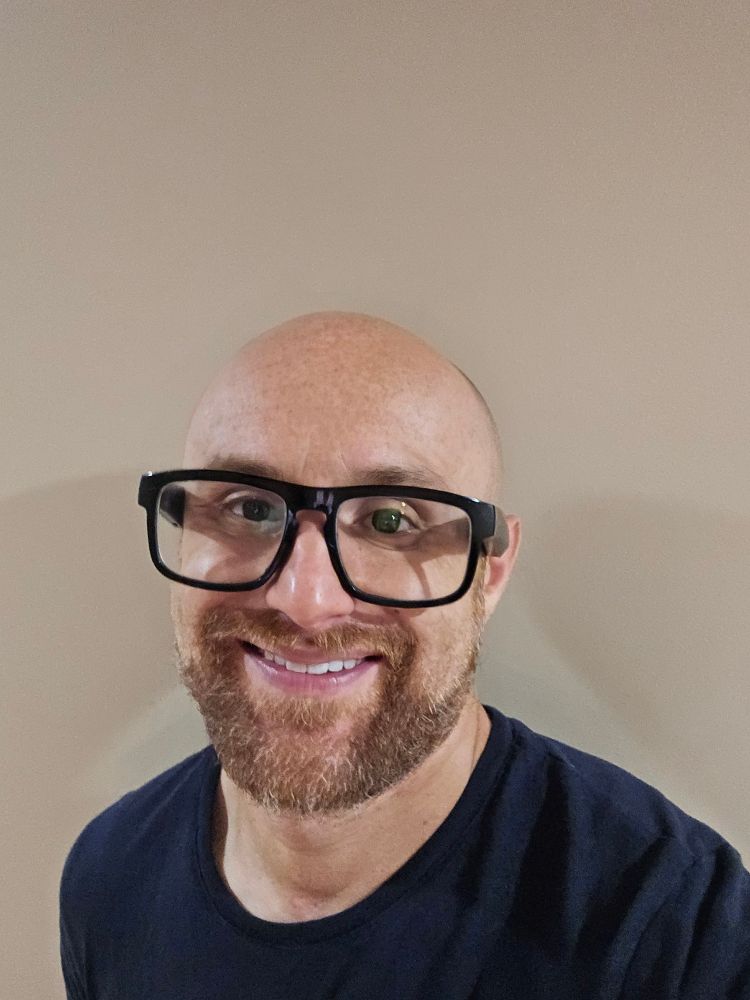 A selfie of me in front of a light brown wall. I'm a bald, middle-aged, white man with a red beard flecked with white. I'm wearing glasses with thick black rims and a dark blue t-shirt.