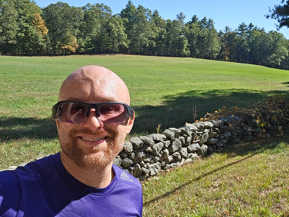 A selfie of me in front of a grass field bordered by a stone wall behind me and forest beyond on a sunny day. I'm a bald, middle-aged, white man with a red beard flecked with white. I'm wearing black sunglasses and a purple adidas running shirt.