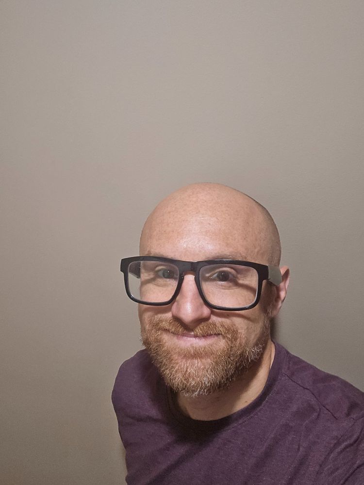 A selfie of me in front of a brown background. I'm a bald, middle-aged, white man with a red beard flecked with white. I'm wearing glasses with thick black rims and a maroon t-shirt.