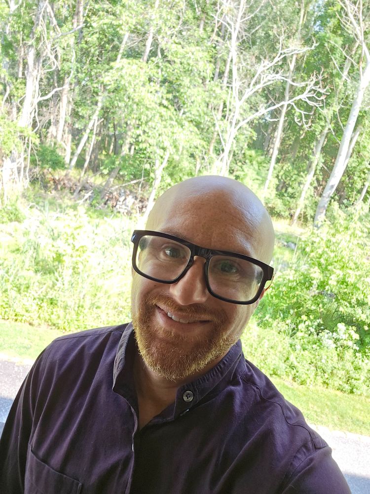 A selfie of me in front of a brightly lit forest. I'm a bald, middle-aged, white man with a red beard flecked with white. I'm wearing glasses with thick black rims and a maroon dress shirt.