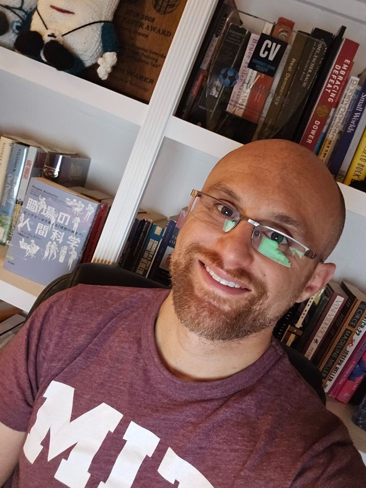 A selfie of me in front of my white bookshelf, which is packed with books. I'm a bald, middle-aged, white man with a red beard flecked with white. I'm wearing glasses with a metal top rim and a maroon t-shirt with MIT written in white across the chest.
