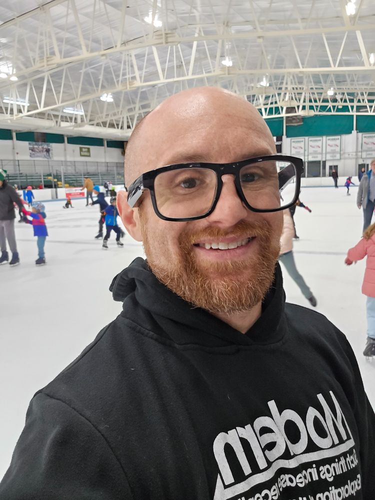 A selfie of me in a brightly lit indoor skating rink. I'm a bald, middle-aged, white man with a red beard flecked with white. I'm wearing thick black-rimmed glasses and a black hooded sweatshirt that says Modern in white letters on it.
