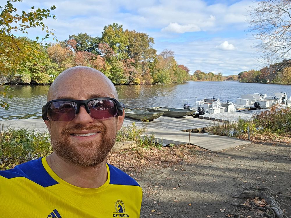 A selfie of me in front of a dock with small boats attached to it on the Charles River on a sunny day. Forest lines both banks, with the leaves mostly green but a few reds and browns as well. I'm a bald, middle-aged, white man with a red beard flecked with white. I'm wearing black sunglasses and a yellow and blue 125th Boston Marathon running shirt.