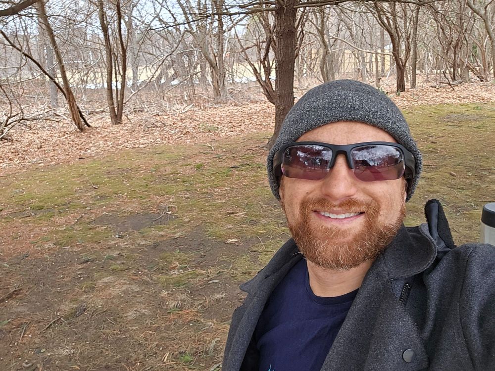 A selfie of me in front of a small forest with some muddy grass in front. I'm a middle-aged white man with a red beard flecked with white wearing a grey winter hat, black sunglasses, and a grey winter coat over a dark blue shirt.