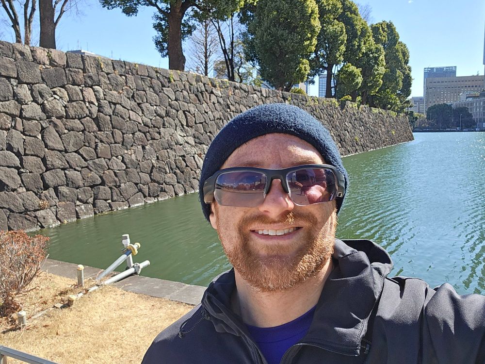 A selfie of me in front of a stone wall of the Japanese Imperial Palace, with trees peaking out on the far side, and a green water moat bordering the wall on a sunny day. I'm a middle-aged white man with a red beard flecked with white. I'm wearing a black headband, black sunglasses, and a black running jacked over a purple shirt.