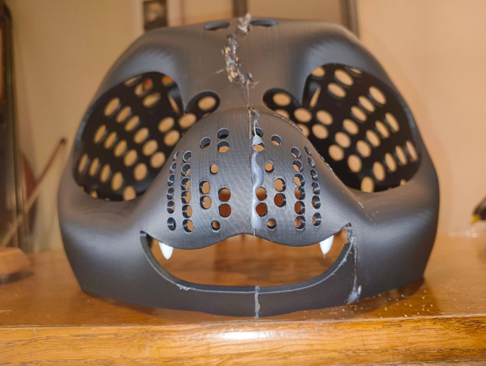 Front close up of a 3d printed fursuit mask, black in colour. It is glued along the center between the eyes, the whole way around from the mouth to the back of the head. 
It also has two white canine teeth