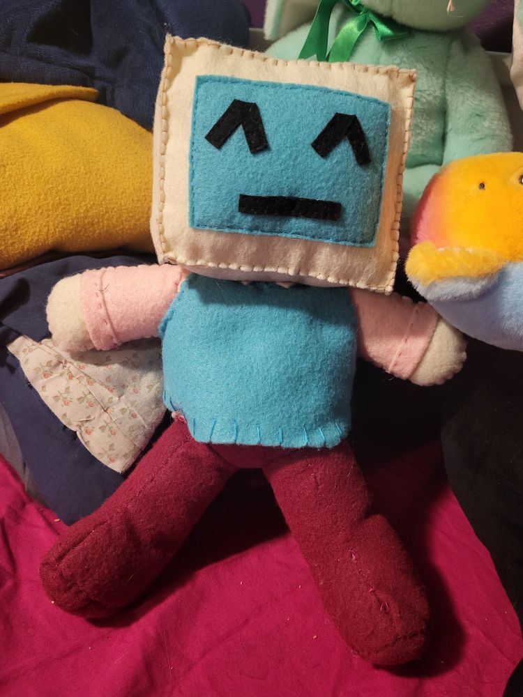 A picture of a monitor head plush. The monitor is beige with a blue screen. On the screen there's a smiling emoticon. The body is chubby. The monitor head is wearing a light pink shirt, a blue sweater vest, and dark pink pants. There is visible sewing on the entirety of the plush.