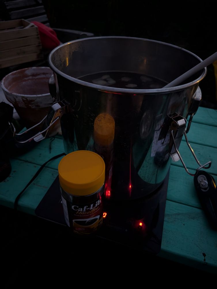 4 gallons of boiling wort on an induction hotplate on a teal picnic table. A half jar of Caf-Lib roasted barley and chicory coffee substitute sits on the edge of the hotplate. There's a can opener and a flower pot in the periphery. It's getting dark.