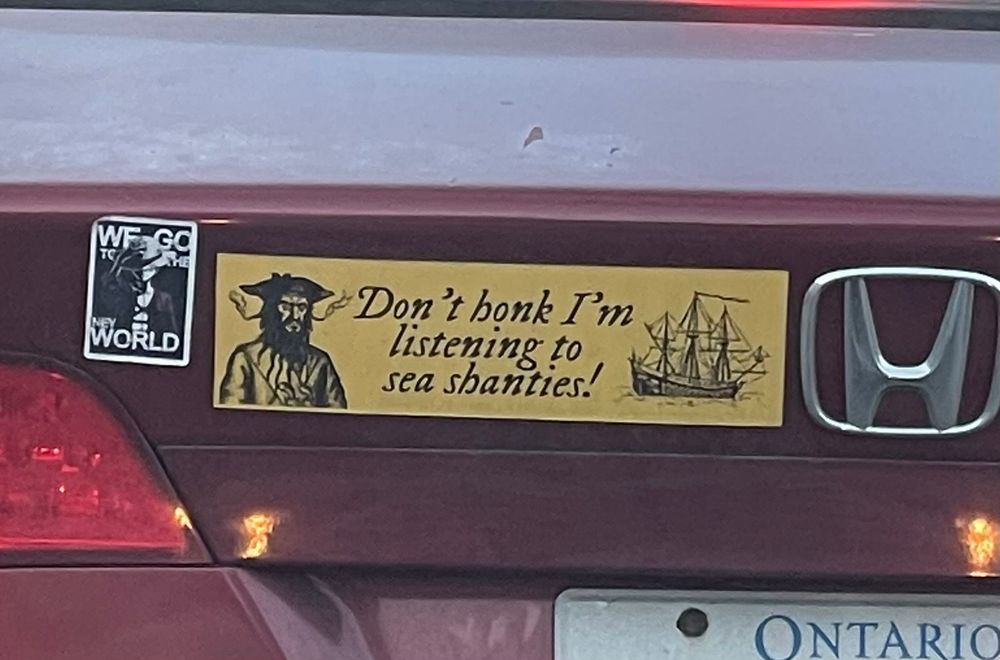 A closeup of a bumper sticker on the back of a burgundy Honda with an Ontario license plate. The sticker is yellow and has the famous illustration of Blackbeard with smoking tapers coming out of his beard on one side and a three-masted ship on the other. In the middle it says “Don’t honk I’m listening to sea shanties!”