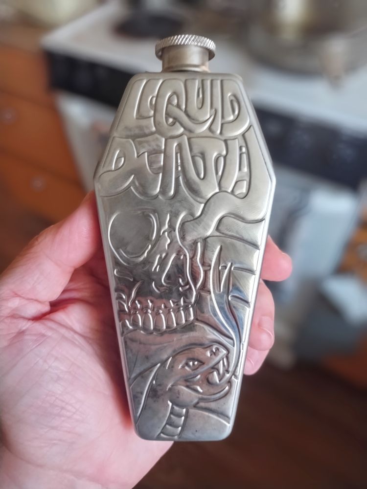 A stainless steel flask shaped like a coffin emblazoned with "liquid death" depicting a kickass skull and snake.