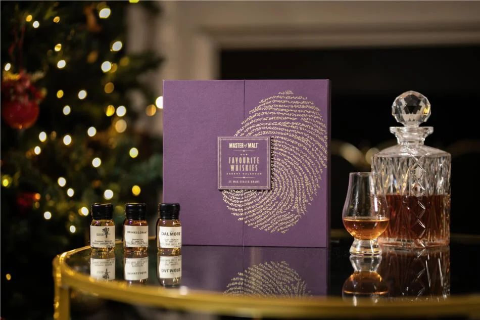 A purple box with a golden fingerprint labeled "Masters of Malt Favourite Whiskies Advent Calendar, 2024 Edition" standing on a glass table in front of a fireplace with a decorated christmas tree out of focus in the background.  3 assorted drams of whiskey stand in front to the left; a decantur and a glencairn glass stand to the right, half filled with whiskey.