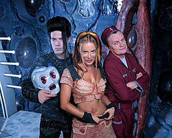 The cast of Lexx: The Series: a tall white goth guy, a robot head, a sexy woman in midriff baring top, a guy who looks like a janitor 