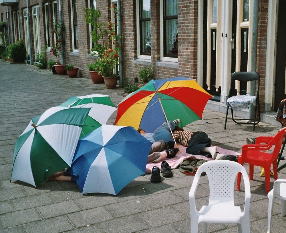 Een straat met stoeptegels waarop vier gekleurde paraplu's die dienen als parasols. Daaronder liggen op een roze deken kinderen te schuilen voor de zon. Rechts zie je enkele (plastic) stoelen verspreid rond het kleed.