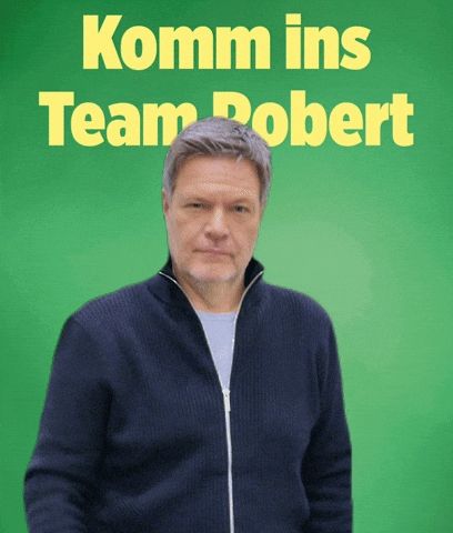 Robert Habeck in einer blauen Strickjacke und grauem T-Shirt drunter steht vor einem grünen Hintergrund auf dem "Komm ins Team Robert" steht. Er streckt seine beiden Arme nach vorn. Seine Daumen sind oben.