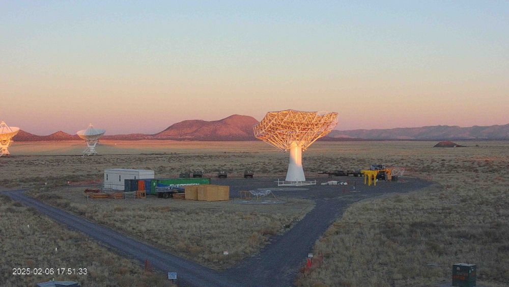 Webcam image showing the ngVLA prototype construction site at sunset. The large white antenna dish is sitting upon its pedestal. In the background, you can see the desert mountains, two VLA dishes, and the colorful sunset skies.