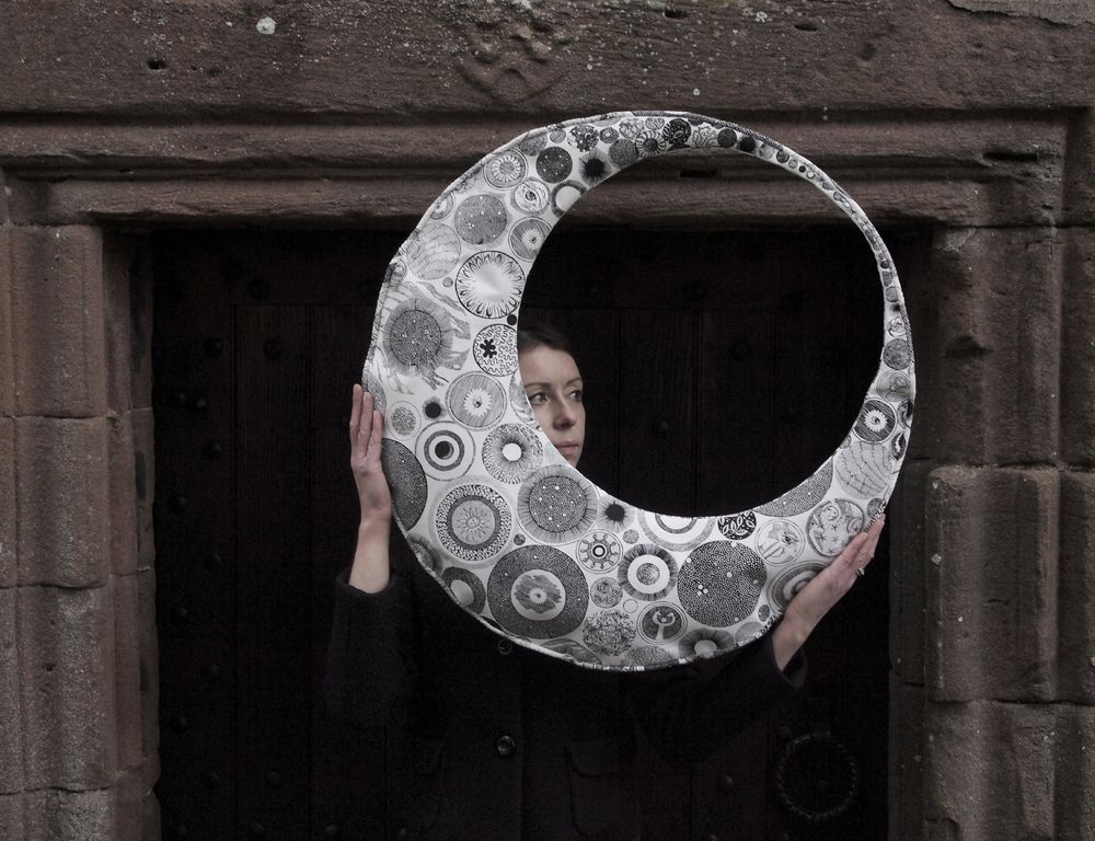 In front of a stone doorway a woman holds a crescent shaped object, patterned in black and white with sun, moon and star forms, her face looking through.