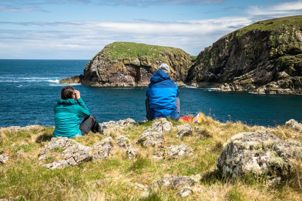 Two people wearing coloured coats, sitting on a cliff overlooking the sea. They are using binoculars to watch birds in the distance. [Photo: Danni Thompson]