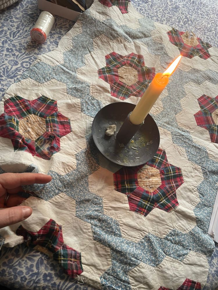 A hand, mending a hand stitched mid nineteenth century hexagon quilt. A candle burns alongside. 