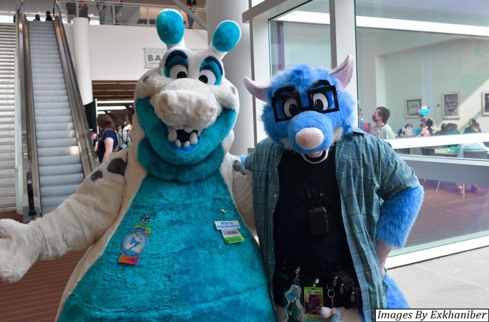 Two fursuiters.  A friendly teal-and-white space alligator and a blue and white mouse stand side-by-side and face the camera.