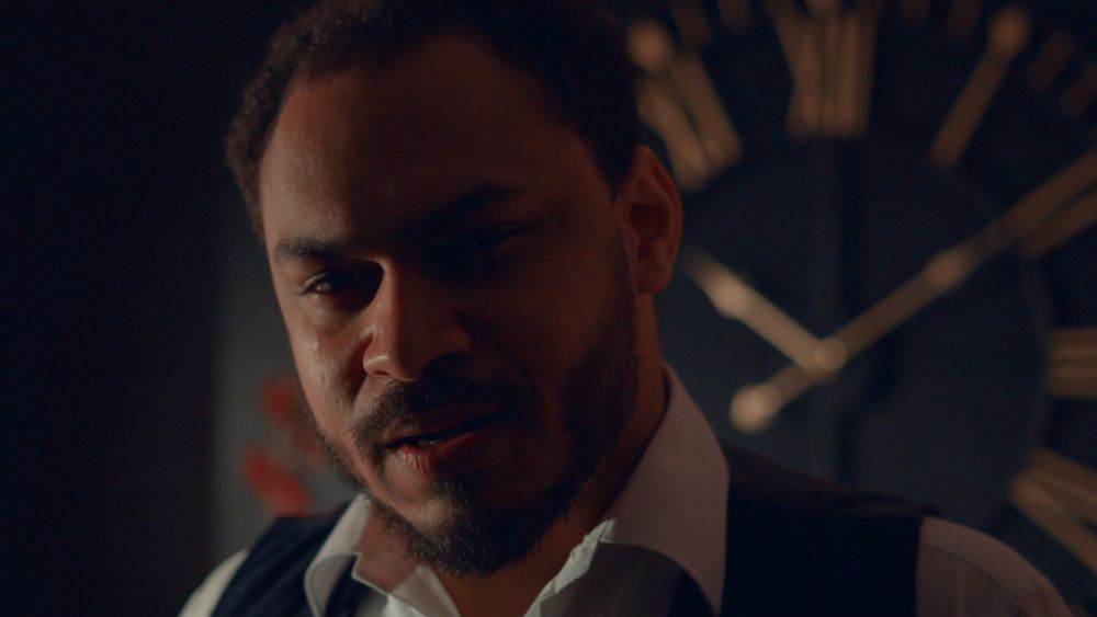 A man in an open-collared white shirt and black waistcoat stands in a dark room. He has an oval face, short dark hair and beard. He looks directly at the viewer with an irate expression. Behind him a large gold clock hangs on the wall. The time is ten past two.
