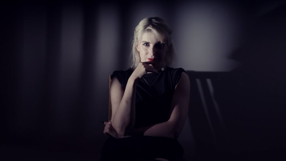 A dark shadowy room. A seated blonde-haired woman in a black sleeveless dress looks directly at the viewer. One arm is folded across her stomach and the other is lifted to her face. Her fingers touch bright red lips which are curled into a knowing smile.