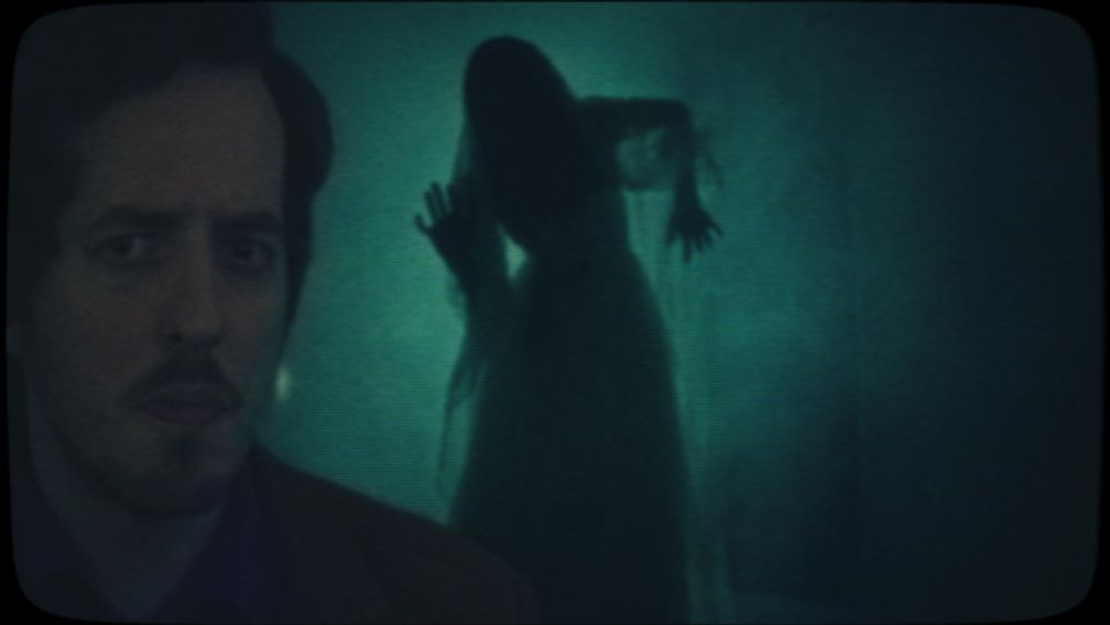 A close up of a curved TV screen. A man with a narrow face, dark hair and beard looms at the left edge of the screen with a serious expression on his face. Behind him, the silhouette of a veiled woman forms a haunting pose against a bluey-green background.