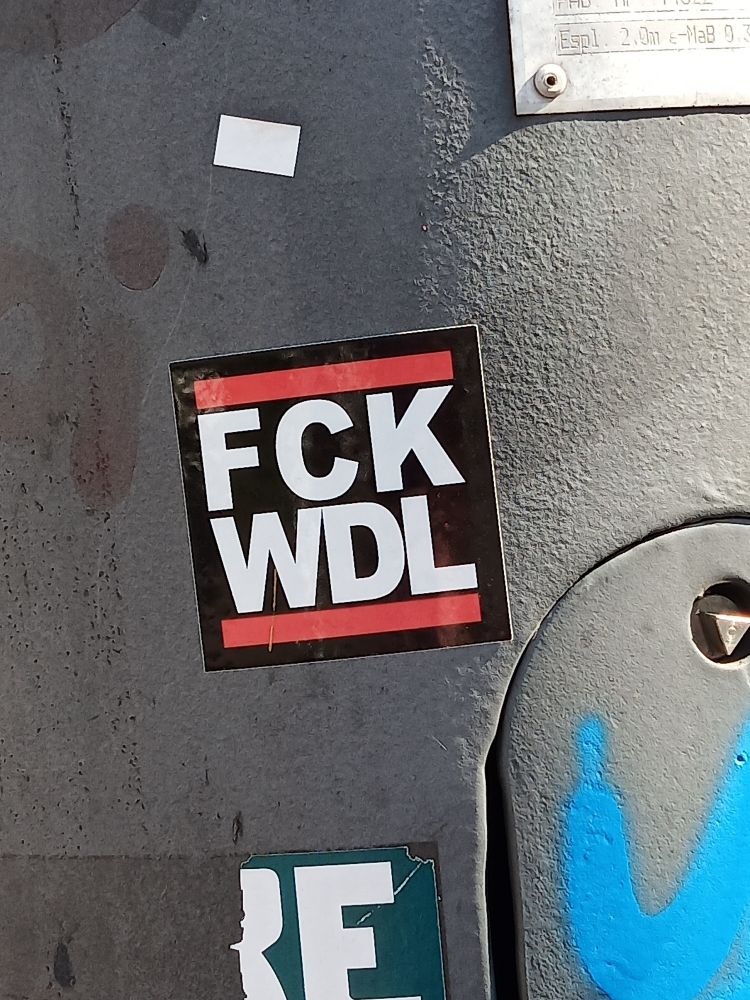 Ausschnitt eines Laternenpfahls, auf dem ein FCK WDL Aufkleber im klassischen FCK NZS Logo klebt 
