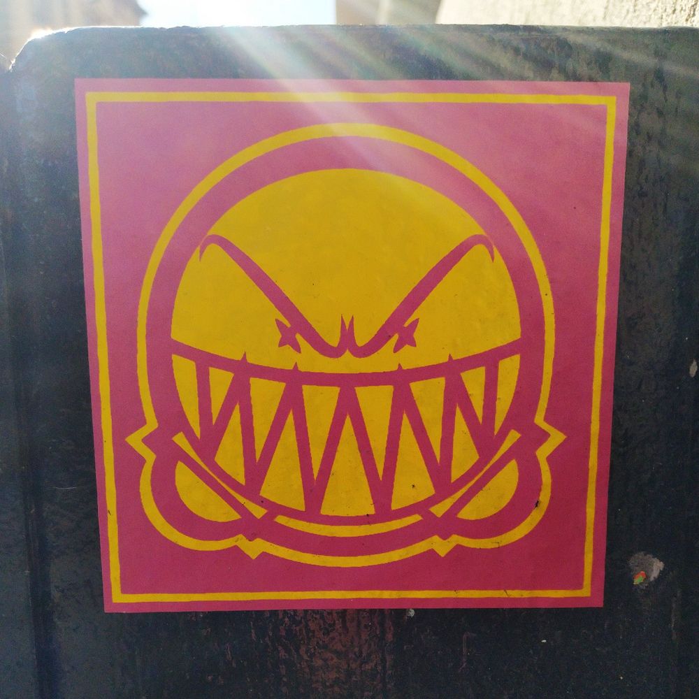 A square sticker about 10cm square. It is a purple-pink with a  highly stylised grinning face rendered in yellow