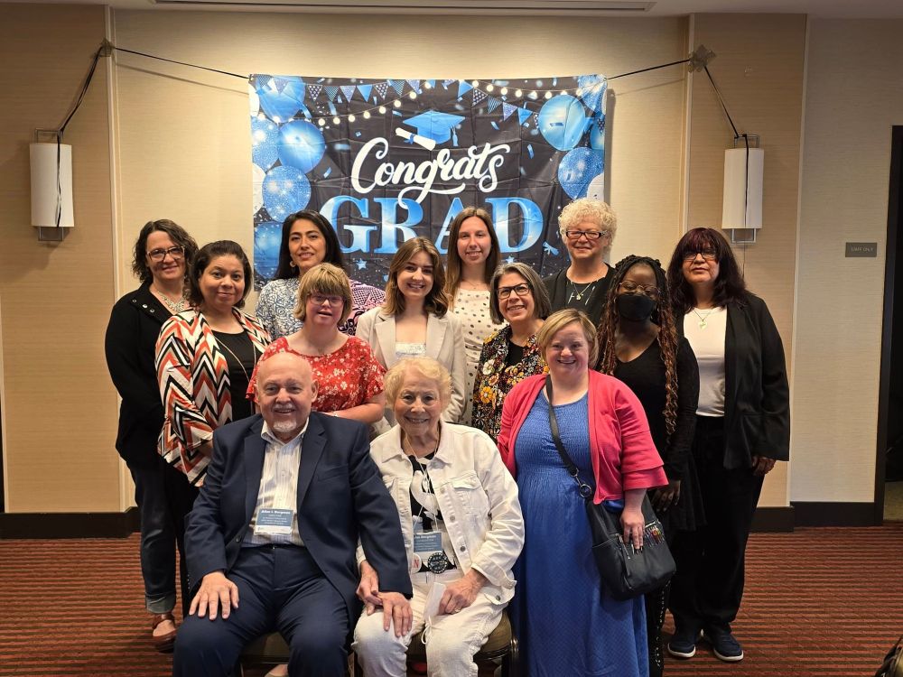 group photo of about 20 people taken in hotel meeting room. Two people seated in front. others are standing behind the two who are seated.