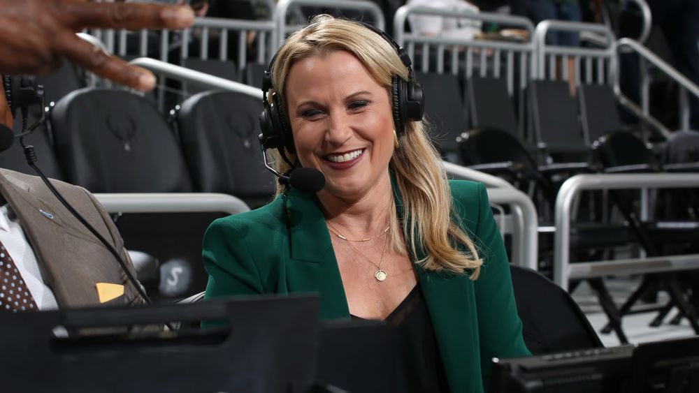 Lisa Byington smiles while wearing a headset microphone during a game broadcast. She has long blonde hair and is dressed in a dark green blazer over a black top, with gold jewelry. Empty black stadium seats and railings are visible in the background.