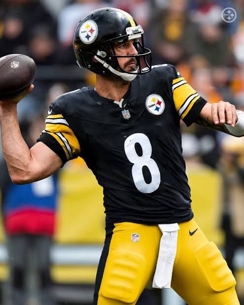 Aaron Rodgers in a Pittsburgh Steelers uniform, number 8, mid-throw during a game. He is wearing a black helmet, black jersey, and yellow pants, holding the football back as he prepares to pass.