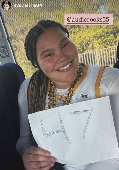 Audi Crooks sits on a bus in her white Iowa State uniform, smiling brightly while holding a handwritten sign that says “47.” She’s wearing two thick gold chains and has long braids. A tag reading “@audicrooks55” appears in the top corner.