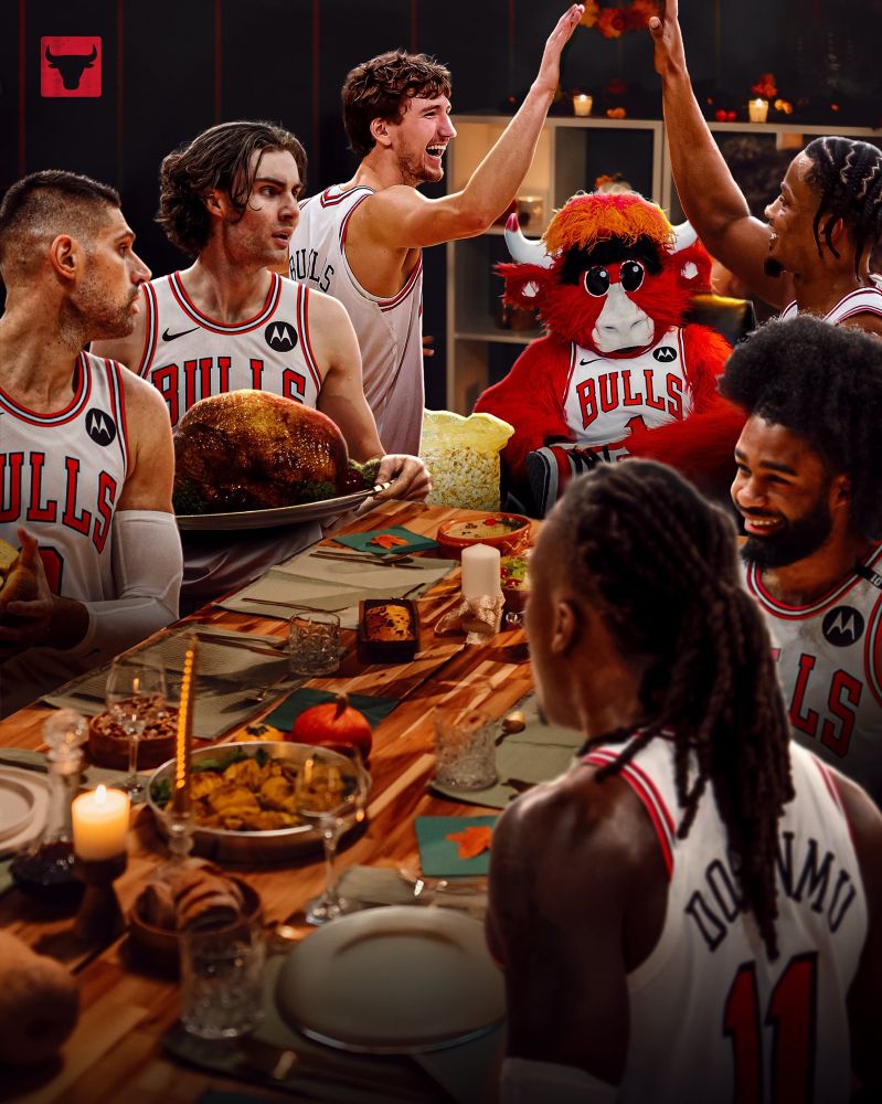 A lively photo of several Chicago Bulls players sharing a Thanksgiving meal at a long wooden table. The players wear white Bulls jerseys and sit among dishes like roasted turkey, bread, and casseroles. Benny the Bull, dressed in a Bulls uniform, is seated in the middle with a huge bowl of popcorn on his lap. Two players high-five above the table while others laugh and talk. Warm, festive lighting fills the room, and pumpkins and candles decorate the scene.