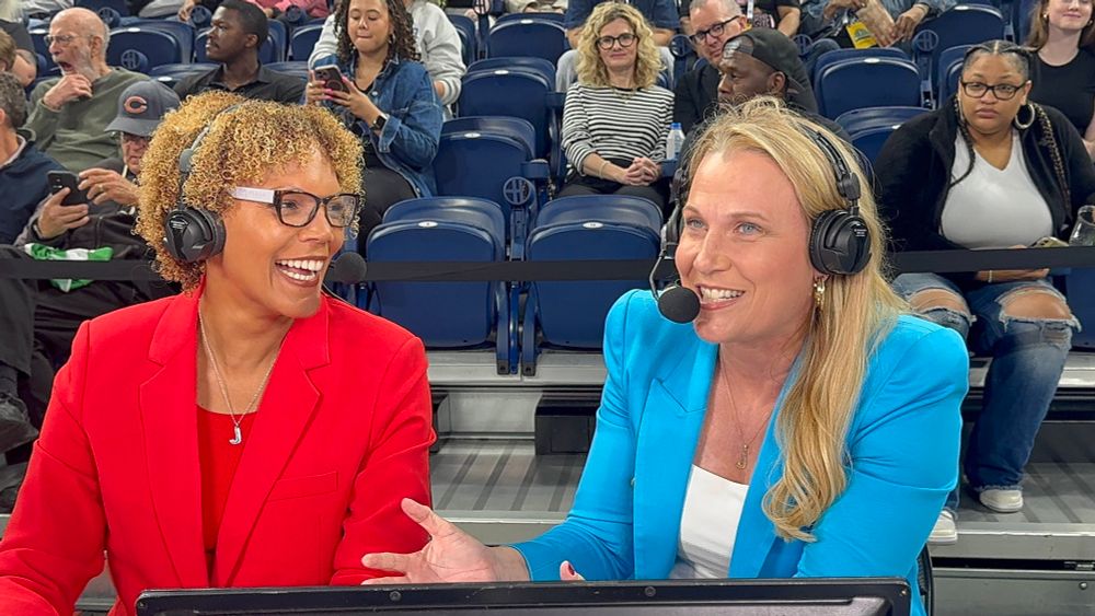 Broadcasters Shimmy Gray-Miller (left) and Lisa Byington (right) sit courtside wearing headsets, smiling and talking during a game. Shimmy wears a bright red blazer, while Lisa wears a light blue blazer. Fans are seated behind them in the stands.