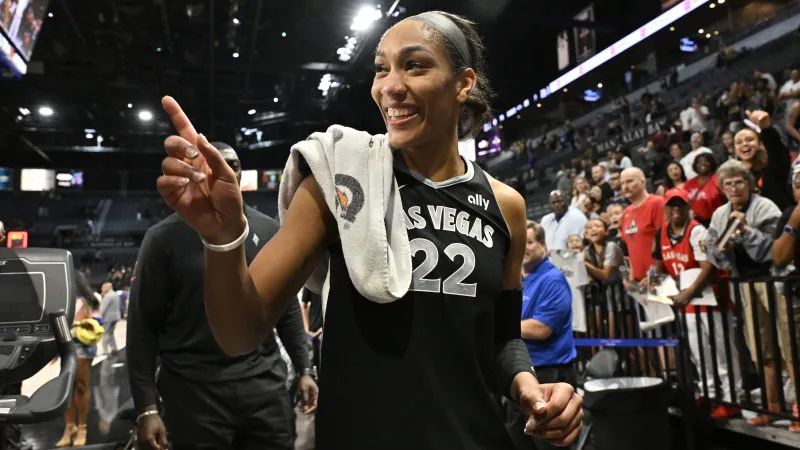 A’ja Wilson, wearing a black Las Vegas Aces jersey with the number 22, smiles and points toward the crowd after a game. She has a towel draped over her shoulder, and fans are cheering in the background.