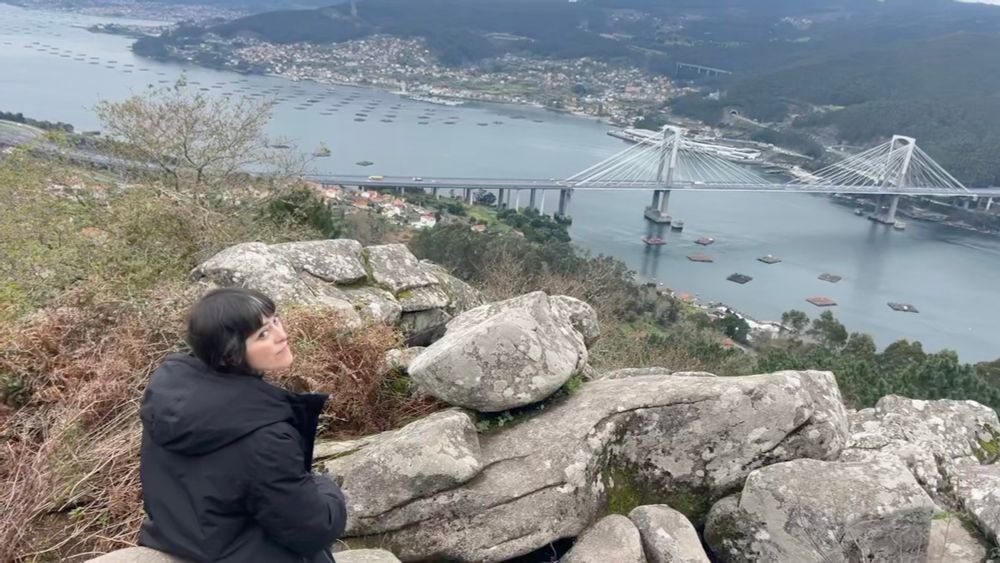 Aparezco sentada en una piedra en lo alto del mirador del campo de la rata en Redondela (Vigo). Estoy mirando a la cámara y detrás de mí aparece el paisaje donde se ve el puente de Rande, la ría, etc.