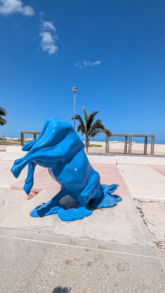 Sculpture of a blue cow melting on the hot pavement on a beach boardwalk. A bright blue sky and palm tree are visible in the background.

