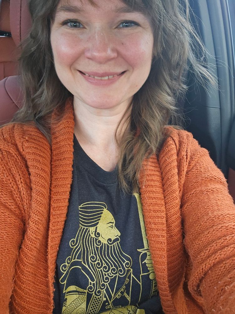 A white woman with brown hair wearing a black ofmd shirt with a gold design of Ed visible, as well as a seasonally-appropriate dark orange cardigan
