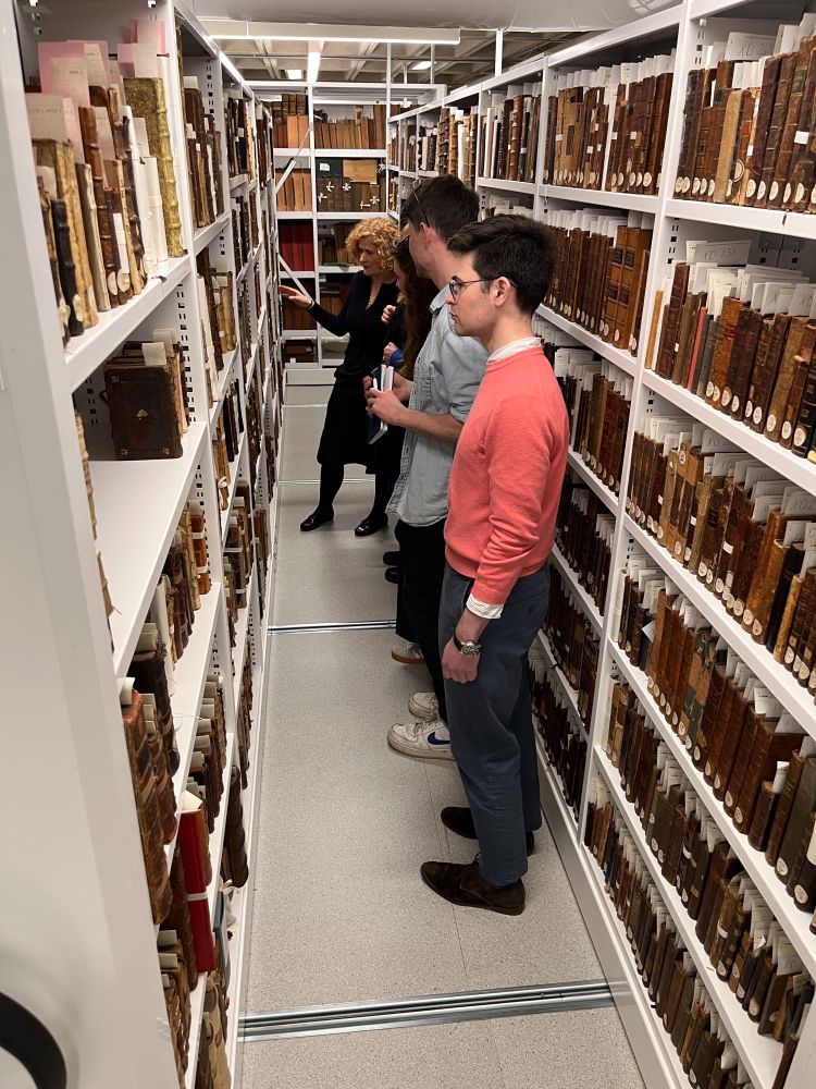 Five people standing looking at old books on white shelves in the storage area of UCD Special Collections.