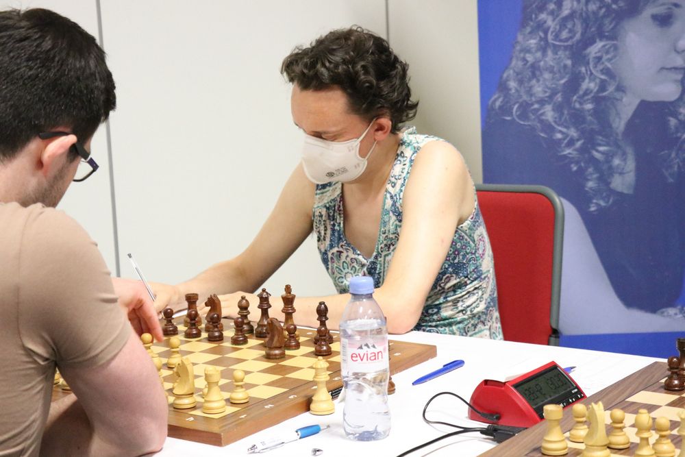 A photo of IM Matthew Wadsworth v IM Jack Rudd from the 2023 British Chess Championships in Leicester. Matthew has his back to the camera; he is a 20-something white man with black hair, wearing a beige t-shirt. Jack, facing the camera, is a 40-something white enby with black hair, wearing a green and white patterned dress and a white face-mask. The position on the board is just after the moves 1.d4 Nf6 2.c4. There is a big poster of IM Jovanka Houska just behind Jack.