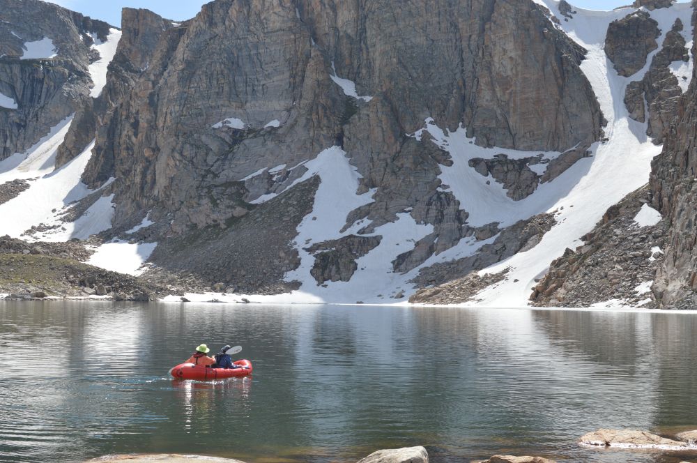 Two scientists in a small red rubber boat paddle across a high alpine lake and are dwarfed by the rocky cliffs on the other side