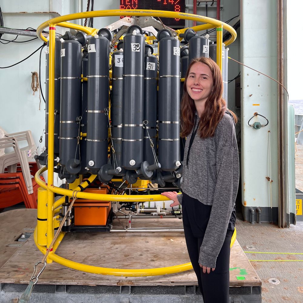 Genevieve Clow standing next to a CTD rosette, a collection of instruments and 36 large water bottles wrapped in a round yellow metal cage. Photo by Roxanne Mina.